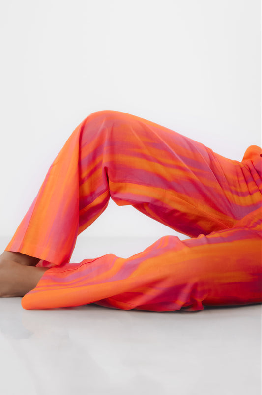 A person reclining on a white studio floor wearing loose, flowing pajama pants in a bright orange, pink, and red abstract striped pattern.
